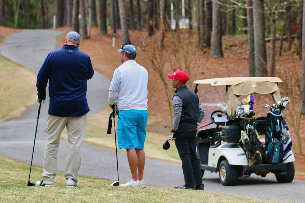 Three men stand by a golf cart holding golf clubs while lookin in the same direction.
