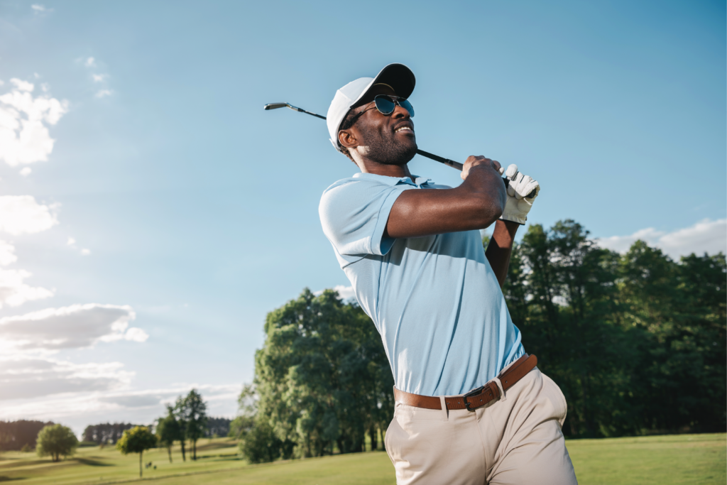 A man in a light blue shirt and white cap is mid swing holding a golf club.